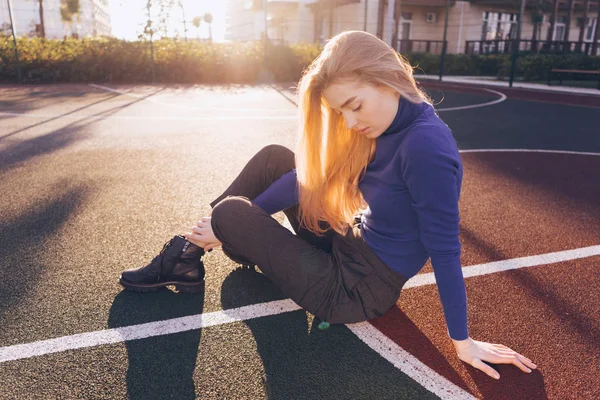 Elegante hermosa chica rubia joven en un suéter azul se sienta en un campo de deportes en el sol —  Fotos de Stock