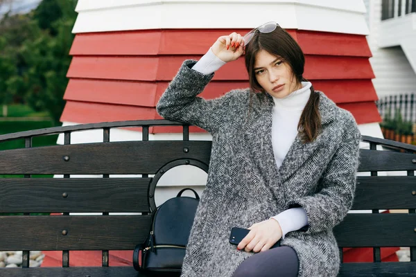 Attractive stylish girl in gray coat sitting on bench, posing — Stock Photo, Image