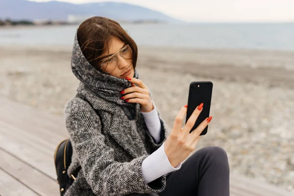 Stylish young girl in a gray coat and glasses walks, makes selfie, cloudy weather — Stock Photo, Image