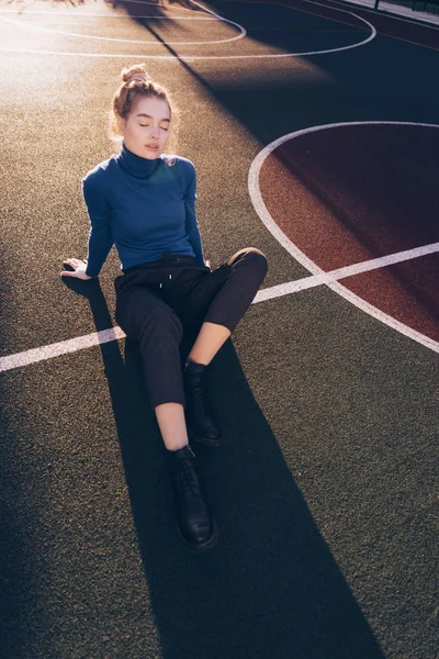 Menina modelo loira bonita elegante em camisola azul elegante senta-se no chão no campo esportivo, posando, fechou os olhos — Fotografia de Stock
