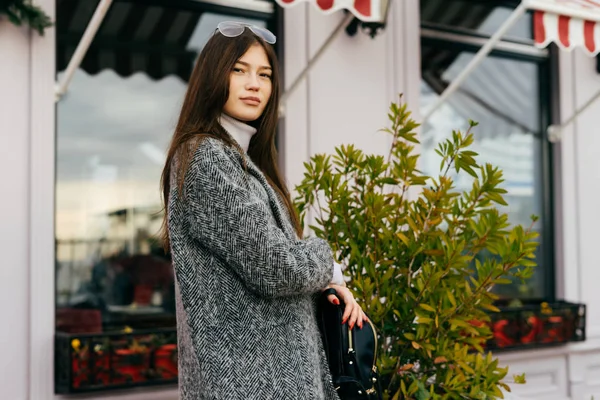 Élégante fille de la ville aux cheveux bruns en manteau gris marchant dans les rues de la ville et posant — Photo