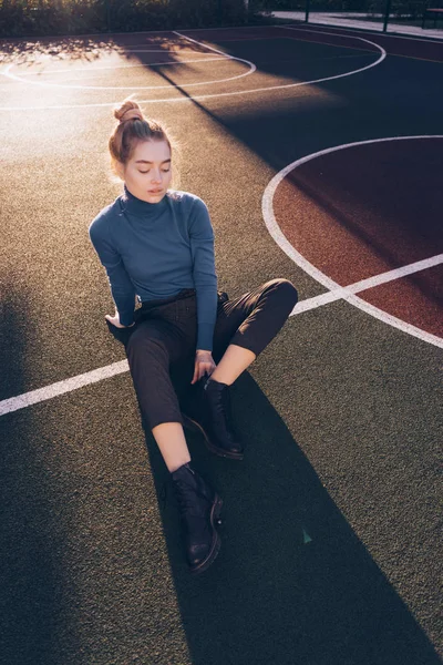 Jonge mooie blonde meisje in stijlvolle kleding zit op een sportveld in de ochtendzon, Streetstyle — Stockfoto