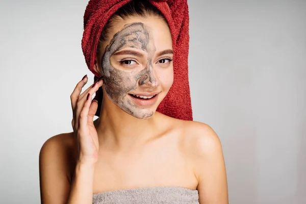 快乐的女孩用毛巾在她的头上应用了一个有用的粘土面具的一半的脸 — 图库照片