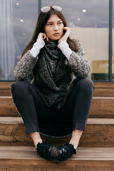 Молодая красивая девушка с темными волосами в очках сидит на ступеньках на улицах города, думает о чем-то — стоковое фото