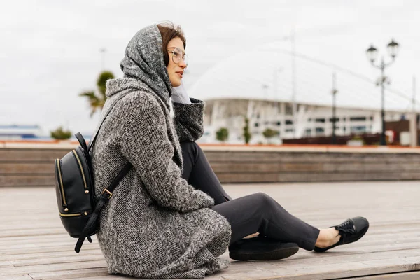 Giovane ragazza alla moda in cappotto grigio e occhiali si siede all'aperto, tempo nuvoloso, stile strada — Foto Stock