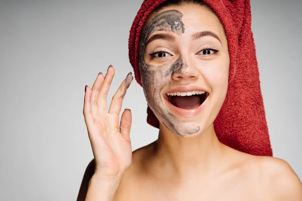 Happy young girl with a red towel on her head applied a clay mask to half of her face, laughs, spa procedures — Stock Photo, Image