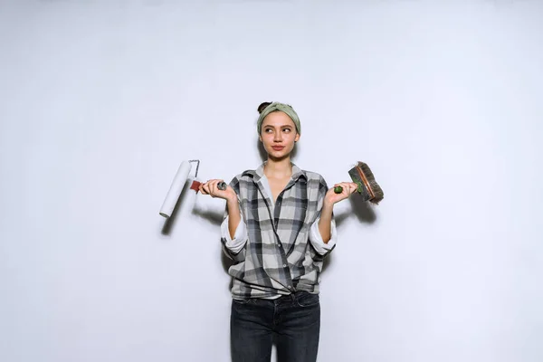 Молодая девушка в клетчатой рубашке делает ремонт в своей квартире, держит кисть и ролик для покраски стены — стоковое фото