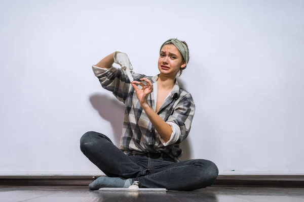 Una joven descontenta en una camisa se sienta en el suelo en su apartamento, se quita los guantes de goma — Foto de Stock