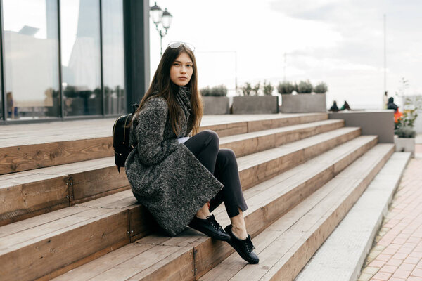 stylish young dark-haired girl in a gray coat sits on the steps in the city streets, posing