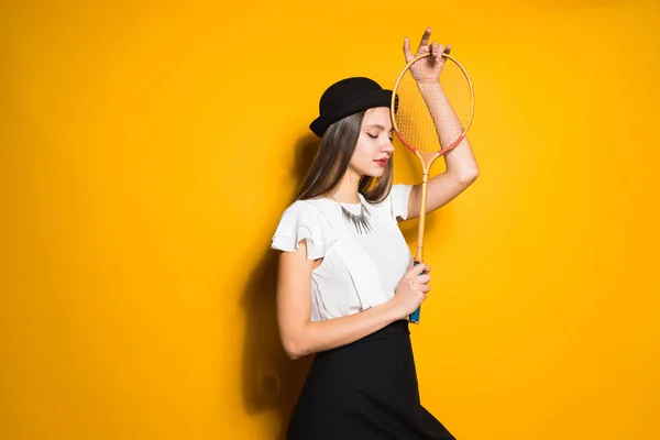 Jovem modelo atraente menina na moda chapéu preto posando no fundo amarelo, segurando uma raquete para jogar tênis — Fotografia de Stock