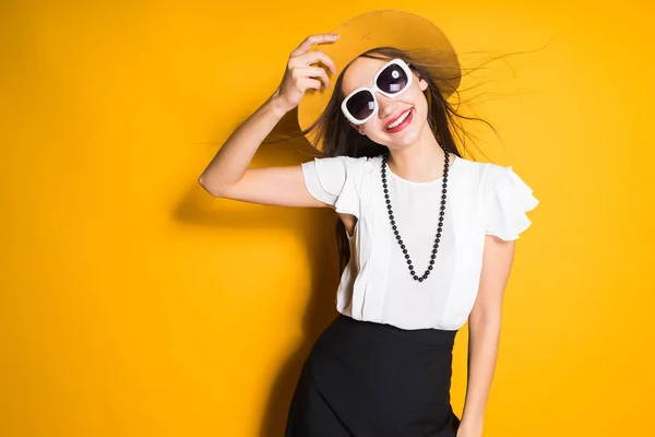 Молодая привлекательная модель девушка в шляпе моды и солнцезащитных очках позирует на желтом фоне, улыбаясь — стоковое фото