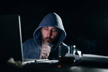 bir adam hacker bir başlıklı bir nargile içiyor, karanlıkta bir dizüstü bilgisayarda yasa dışı bir şey yok