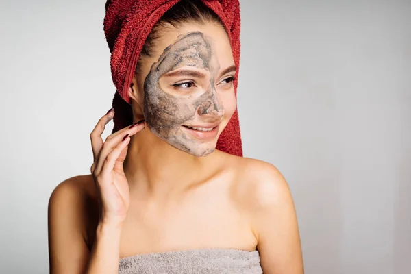 Happy young girl with a red towel on her head puts a useful mask on the face, wants to be beautiful, smiles — Stock Photo, Image
