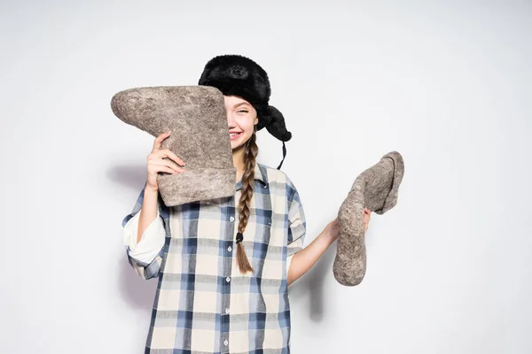 Riendo joven rusa chica en un sombrero con orejeras sostiene gris fieltro botas en las manos — Foto de Stock