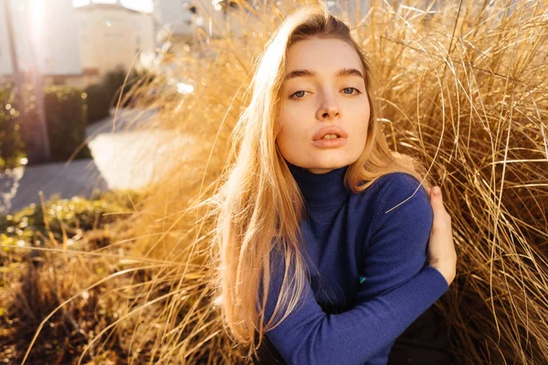 Attractive young blond model girl in blue sweater sits on the background of a yellow plant posing in the sun — Stock Photo, Image