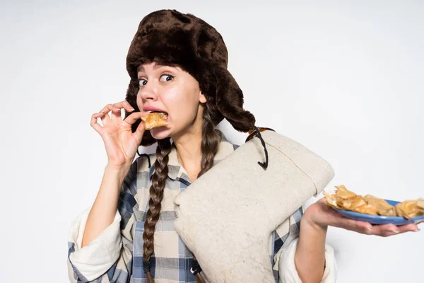 Lustiges russisches Mädchen mit warmem Hut und Ohrenklappen feiert Fastnacht und isst eifrig heiße Pfannkuchen — Stockfoto