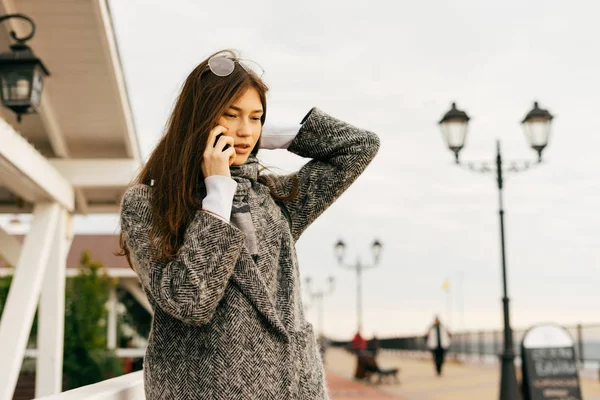 Giovane ragazza di città elegante in cappotto grigio parlando al telefono, passeggiando per la città — Foto Stock