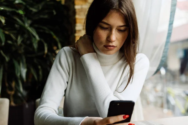 Young dark-haired girl in a white sweater sits in a cafe, waits for food and looks in her smartphone — Stock Photo, Image