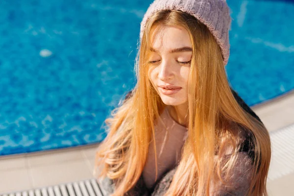 Мила молода дівчина з світлим волоссям, в рожевому капелюсі сидить біля блакитного басейну, на сонці — стокове фото
