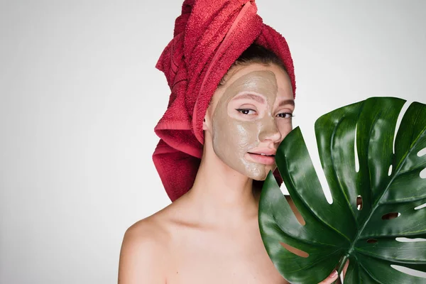 Ένα νεαρό κορίτσι με μια κόκκινη πετσέτα στο κεφάλι της εφαρμόζεται μια χρήσιμη άργιλος μάσκα στο πρόσωπό της, κατέχει ένα πράσινο φύλλο, ημερήσιο σπα — Φωτογραφία Αρχείου