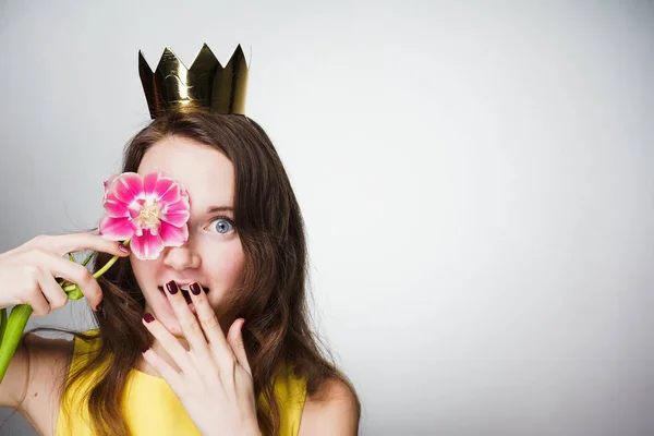 Joven sorprendida en un vestido amarillo sostiene una flor rosa, en su cabeza una corona de oro, celebra el día de la madre — Foto de Stock