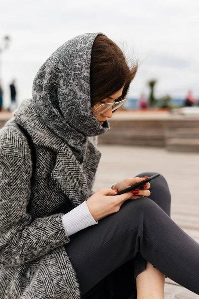 Nice dark-haired girl in a gray coat and glasses writes a message on smartone — Stock Photo, Image