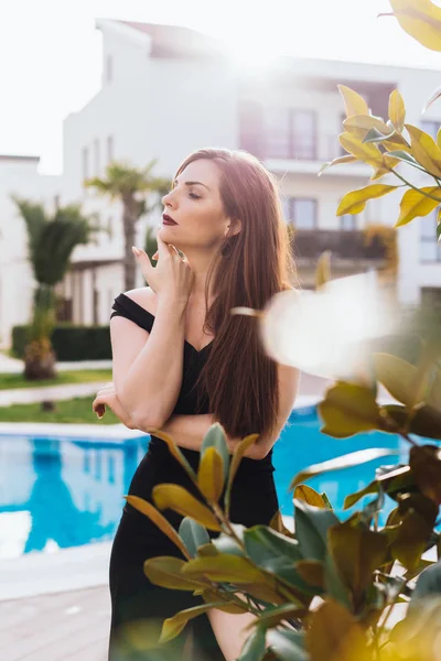 Atraente sexy menina de cabelos longos em um vestido preto goza de uma vida luxuosa e rica, à beira da piscina — Fotografia de Stock
