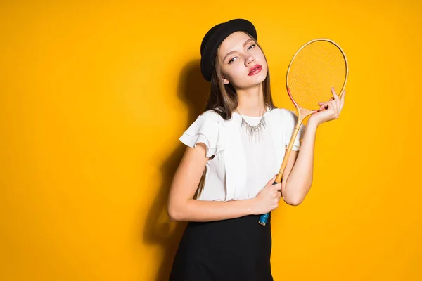 Aantrekkelijk meisje in een zwarte hoed poseren op een gele achtergrond, houden een tennisracket — Stockfoto