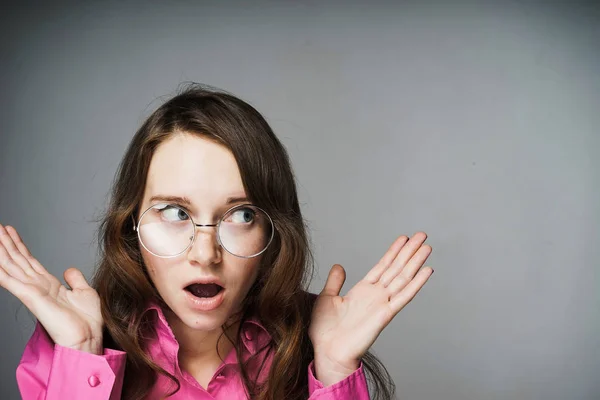 Молодая женщина-офисный работник в розовой рубашке и очках удивлена своими руками, не знает ответа — стоковое фото