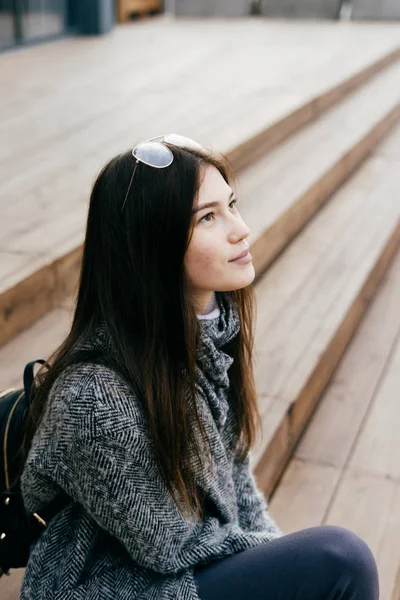 Giovane ragazza dai capelli scuri adolescente in cappotto grigio si siede su gradini e alza lo sguardo al cielo — Foto Stock