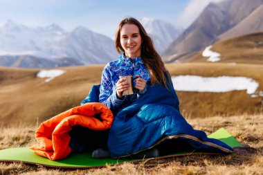 mavi bir ceket ve mutlu aktif genç kız arka planı Kafkas Dağları'nın uyku tulumu içinde oturur, içecekler sıcak çay ve gülümsüyor