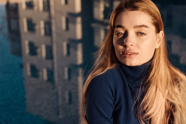 Mooie jonge blonde model meisje in blauwe trui zit bij een blauwe zwembad poseren in de zon — Stockfoto