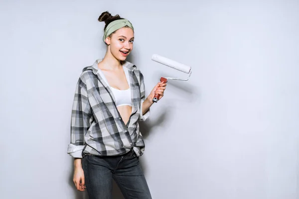 Счастливая молодая девушка в большой клетчатой рубашке держит ролик для покраски стен, ремонт в квартире — стоковое фото