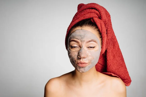 Μια έκπληξη νεαρή κοπέλα με μια κόκκινη πετσέτα στο κεφάλι της εφαρμόζεται μια χρήσιμη μάσκα στο πρόσωπό της, απολαμβάνει θεραπείες spa — Φωτογραφία Αρχείου