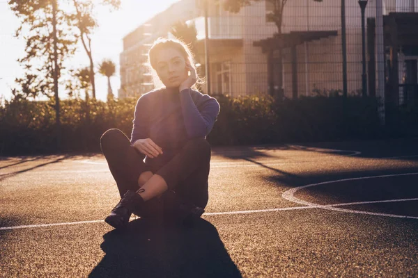 Menina loira nova elegante em um suéter azul senta-se em um campo de esportes nos raios do sol da noite — Fotografia de Stock