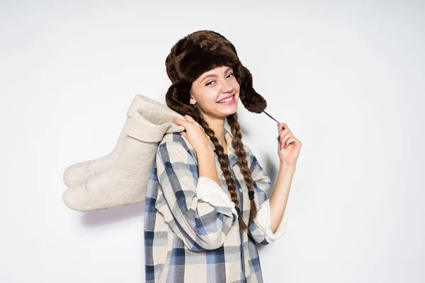 A smiling young girl from Russia in a warm fur hat holds gray felt boots in her hands, her hair is braided — Stock Photo, Image