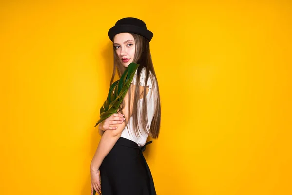 Chica atractiva con confianza en sombrero negro de moda posando sobre fondo amarillo, sosteniendo una hoja verde — Foto de Stock