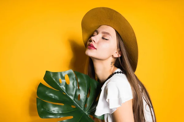 Attractive young girl in fashion hat posing on yellow background, holding green leaf — Stock Photo, Image