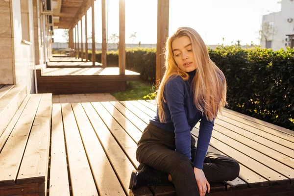 एक ट्रेंडी ब्लू स्वेटर में सुंदर युवा सुनहरे बालों वाली लड़की सूरज में बाहर बैठे — स्टॉक फ़ोटो, इमेज