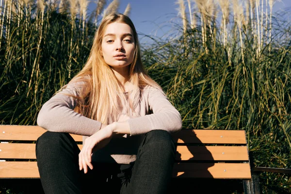 Menina loira atraente confiante sentado em um banco ao ar livre, em um parque, apreciando o sol e calor — Fotografia de Stock