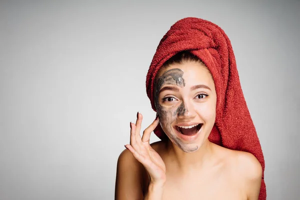 Happy young girl with a red towel on her head puts a clay mask on half the face — Stock Photo, Image