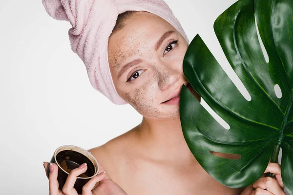 Hermosa joven con una toalla rosa en la cabeza pone un exfoliante de café en su cara, sostiene una hoja verde, spa de día —  Fotos de Stock