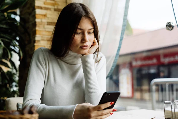 Beautiful dark-haired girl in a white sweater sits in a cafe and looks in a smartphone — Stock Photo, Image