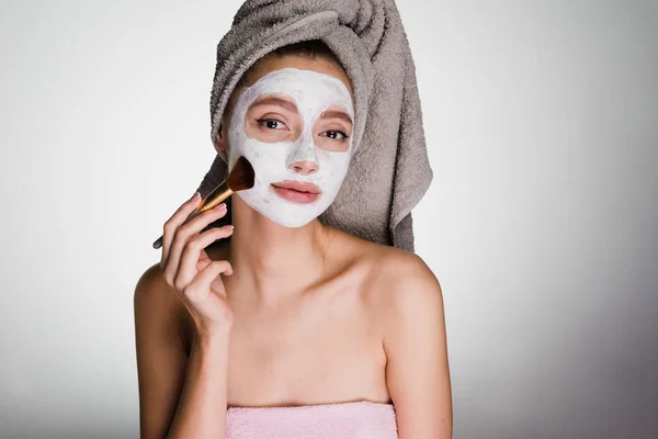 Linda joven con una toalla en la cabeza se pone una máscara hidratante blanca en la cara, usando un cepillo — Foto de Stock