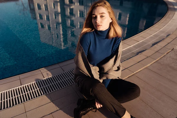 Bela menina loira atraente em camisola azul senta-se à beira da piscina azul, gosta do sol da primavera e olha para a câmera — Fotografia de Stock