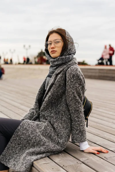 Стильна молода міська дівчина в сірому пальто і окулярах сидить біля моря, дивлячись на камеру — стокове фото