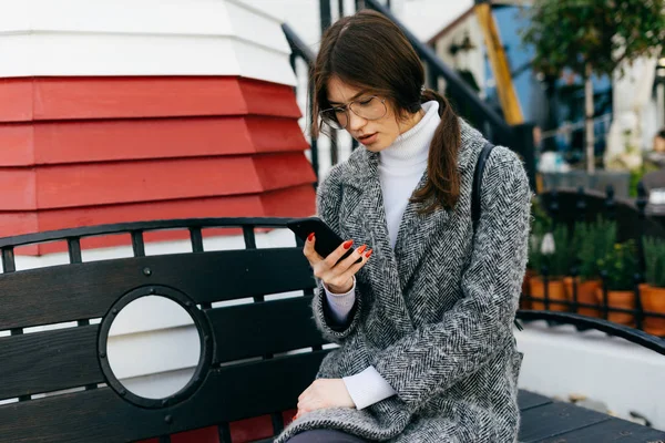 Stylish young dark-haired girl with glasses and a gray coat writes a message on the smartphone — Stock Photo, Image