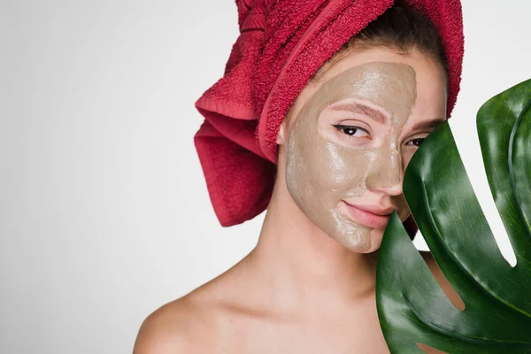 A cute young girl enjoys a spa, has put a clay mask on half of her face, holds a green leaf — Stock Photo, Image