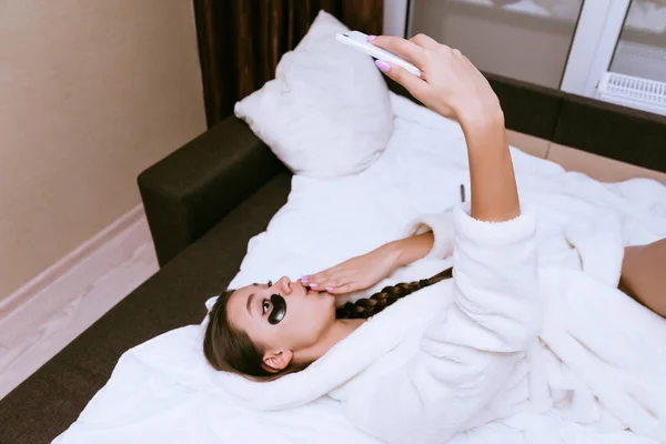 Забавная девушка в белом халате лежит на кровати, под глазами черных пятен, делает селфи — стоковое фото