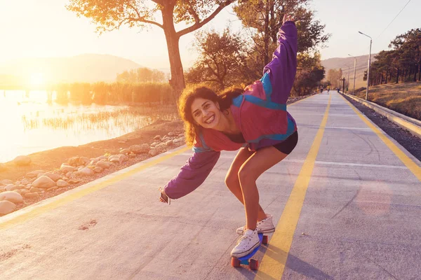 Felice ragazza ridente in una giacca alla moda sta cavalcando un longboard sulla strada al tramonto — Foto Stock